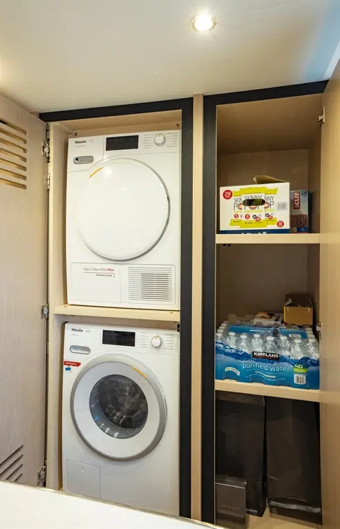 Miele washer and dryer stacked in a wooden cabinet, with boxes of popcorn and bottles of purified water visible above.